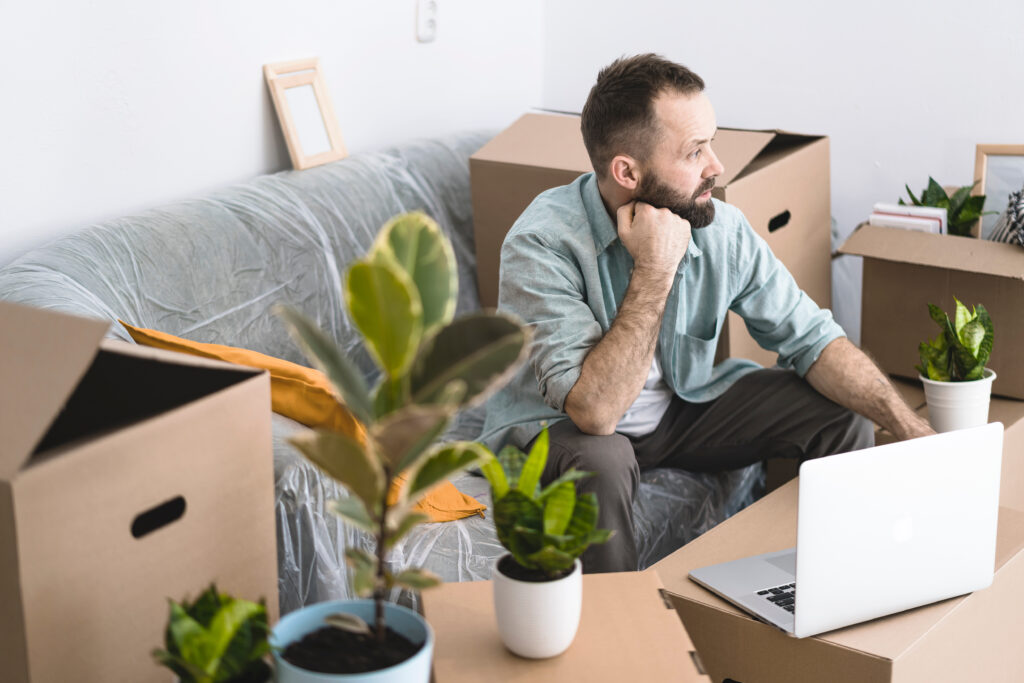 A mature man sitting in a messy room with cardboard boxes and plants, using laptop.