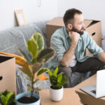 A mature man sitting in a messy room with cardboard boxes and plants, using laptop.