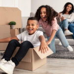 Portrait of happy black family playing and celebrating relocation, laughing daughter riding her brother in carton container, parents hugging while sitting on couch in background